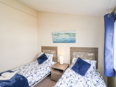 A bedroom with two single beds, a bedside table with a lamp, a painting above the beds, and a window with blue curtains at Two Little Ducks in Cayton