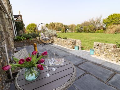 A patio with a round table holding flowers wine glasses and a bottle a hanging chair and a stone wall with grass beyond at The Roost in Porthallow