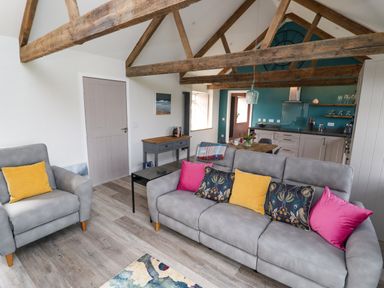 A living room with gray sofas and colorful cushions next to a kitchen with teal walls and wooden beams at The Roost in Porthallow
