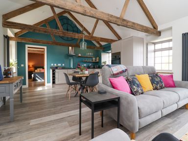 An open living space with exposed wooden beams a grey couch with colorful cushions a dining table with black chairs and a kitchen area with teal walls at The Roost in Porthallow