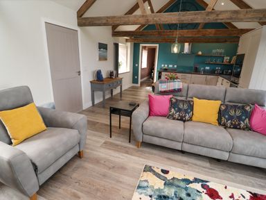 A living room with gray sofas and colorful cushions next to a kitchen with wooden beams at The Roost in Porthallow