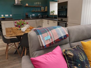 A kitchen and dining area with a wooden table set with cups and flowers and a sofa with colorful cushions and a blanket at The Roost in Porthallow