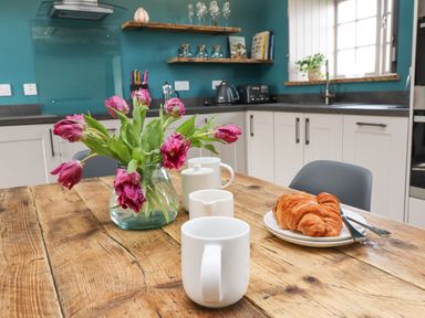 A kitchen with white cabinets a wooden table with pink flowers white mugs and croissants at The Roost in Porthallow