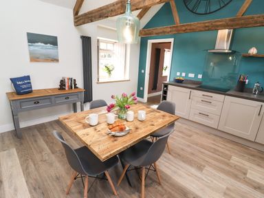 A kitchen dining area with wooden beams wooden table gray chairs flowers on table white mugs croissants painting and cabinets at The Roost in Porthallow