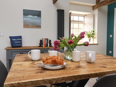 A dining table with four white mugs croissants and flowers in a vase in a room with a window and a sideboard at The Roost in Porthallow