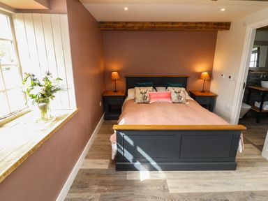 A bedroom with a double bed pillows and bedside tables with lamps and a vase of flowers on the window sill at The Roost in Porthallow