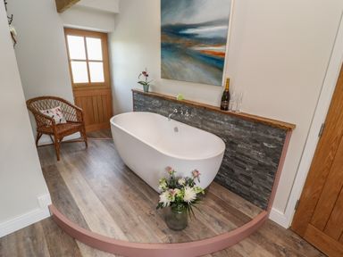 A bathroom with a freestanding tub a wicker chair and a door at The Roost in Porthallow