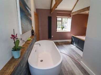 A bathroom with a freestanding bathtub orchid on the ledge and a bedroom with a bed visible through an open space at The Roost in Porthallow