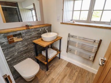 A bathroom with a wall-mounted toilet wooden sink stand with a white basin and folded towels a towel radiator and a window at The Roost in Porthallow