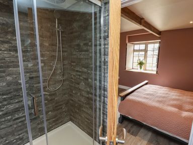 A bedroom with a bed covered in a quilt facing a window and an adjacent bathroom with a glass shower enclosure at The Roost in Porthallow