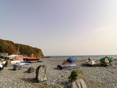 A pebbled beach with several small boats on the shore and a rocky hill in the background at The Roost in Porthallow