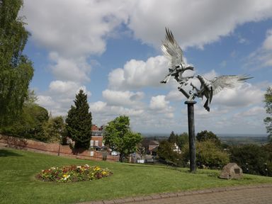 A park with a metal bird sculpture a flower bed and trees with buildings in the background at The Escape in Malvern