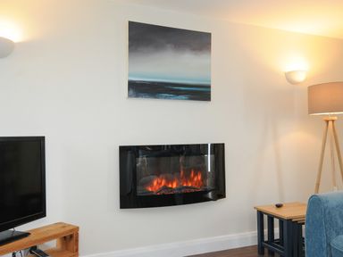A room with a wall-mounted electric fireplace a television on a wooden stand a painting above the fireplace a wooden floor lamp and a small wooden table at The Bower in Manaccan near St Keverne