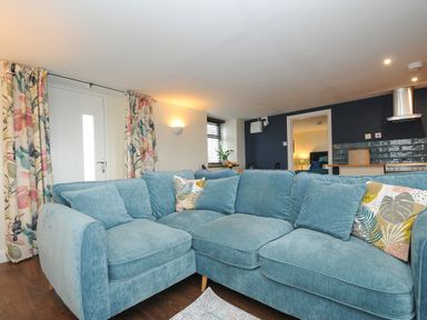 A living room with a blue sectional sofa cushions floral curtains and a kitchen area at The Bower in Manaccan near St Keverne