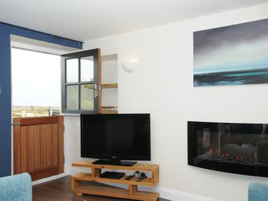 A corner of a living room with a wooden door open above blue wall section a flat screen tv on a wooden stand and an electric fireplace on a white wall at The Bower in Manaccan near St Keverne