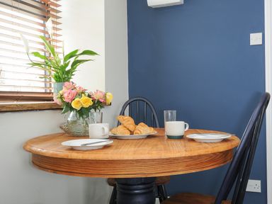A wooden round dining table with two chairs plates cups croissants and flowers next to a window at The Bower in Manaccan near St Keverne