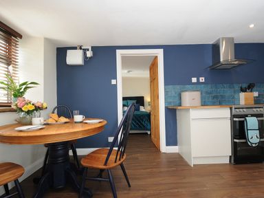 A kitchen with a wooden table set for breakfast and a stove next to a doorway showing a bedroom at The Bower in Manaccan near St Keverne
