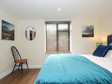 A bedroom with a bed covered in teal bedding a wooden chair a round mirror two paintings and a window with wooden blinds at The Bower in Manaccan near St Keverne