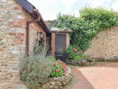 A stone cottage exterior with a wooden door and a small patio area with plants and flowers at Coach Cottage in Colyton