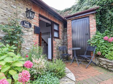 An outdoor seating area with two black chairs and a small round table next to a wooden door with plants and flowers outside at Coach Cottage in Colyton