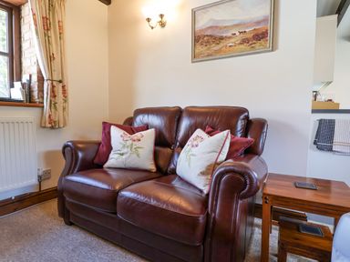 A leather sofa with floral and plain cushions next to a wooden side table under a wall painting at Coach Cottage in Colyton