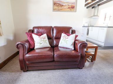 A brown leather sofa with floral and red cushions in a living room next to nested wooden tables and a kitchen in the background at Coach Cottage in Colyton