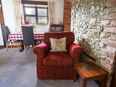 A red armchair with a floral cushion next to a wooden side table in a room with a stone wall and a dining table with chairs at Coach Cottage in Colyton