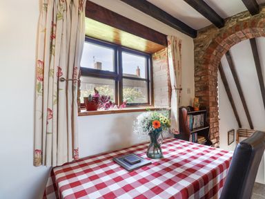 A room with a table covered with a red and white checkered tablecloth and a vase of flowers on it next to a window with floral curtains and a brick archway with a bookshelf at Coach Cottage in Colyton