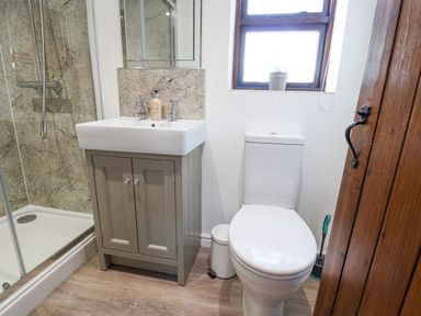 A bathroom with a shower, sink with cabinet, toilet, small trash bin, and a window at Coach Cottage in Colyton