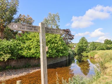 A wooden signpost by a river with green trees and a blue sky at Coach Cottage in Colyton