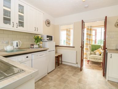 A kitchen with white cabinets appliances a microwave and a view into a sunroom with an armchair at Doves Nest in Martham