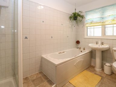 A bathroom with a bathtub, sink, toilet, yellow rug, hanging plant, shower area, and window with seaside-themed blind at Dove's Nest in Martham