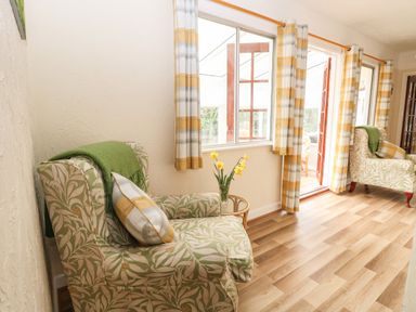 A sitting area with two patterned armchairs a small glass table with flowers and large windows with yellow and white curtains at Doves Nest in Martham