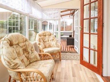 A sunroom with two cushioned wicker chairs open door to a patio with wicker furniture at Doves Nest in Martham