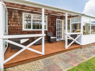 A wooden deck with three brown wicker chairs with white cushions outside a house with a glass door and windows at Dove's Nest in Martham