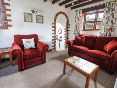 A living room with red armchair and sofa a wooden coffee table and floral curtains at Stable Cottage in Colyton