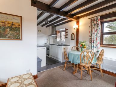 A dining area with a round table and four wooden chairs next to a window and an open kitchen with white cabinets at Stable Cottage in Colyton