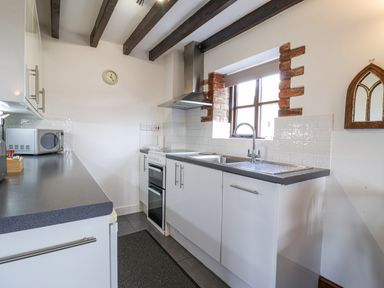 A kitchen with white cabinets a double sink stove microwave and window with exposed brick at Stable Cottage in Colyton