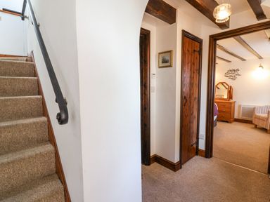 A hallway with carpeted stairs and wooden doors leading to a room with a wooden dresser and striped chair at Stable Cottage in Colyton