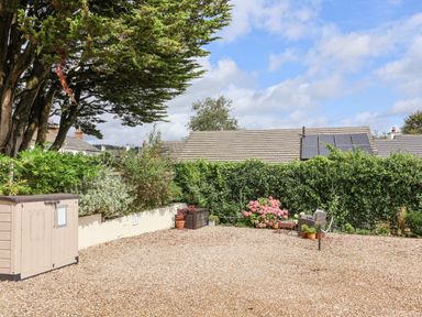 A gravel yard with potted plants a storage box and a chair with a table near a hedge at Stable Cottage in Colyton