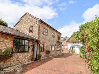 A stone cottage along a brick driveway with plants and a thatched roof house in the background at Stable Cottage in Colyton