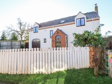 A white house with brown windows and doors behind a white wooden fence with grass in front at Anvil Cottage in Appleton Wiske