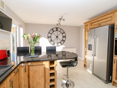 A kitchen with wooden cabinets black countertop bar stools large wall clock and a stainless steel refrigerator at Anvil Cottage in Appleton Wiske