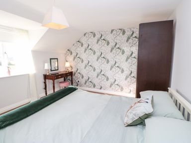 A bedroom with a bed pillow, a green blanket, a dark brown wardrobe, a wooden dressing table with lamps and a mirror, and wallpaper with peacock design at Anvil Cottage in Appleton Wiske
