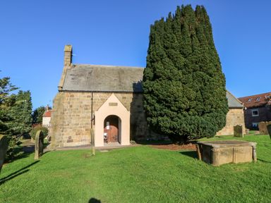 A stone building with an arched wooden door next to a large tree and gravestones on a grassy area at Anvil Cottage in Appleton Wiske