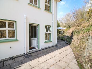 An exterior view of a white house with green window frames and an open door next to a paved pathway leading to a garden area with a picnic table and umbrella at 1 Closheen Lane in Rosscarbery County Cork