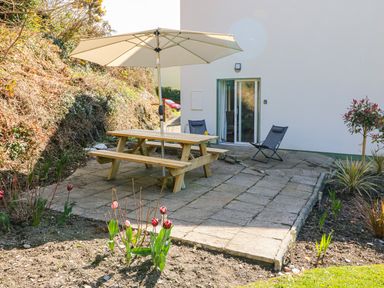 An outdoor patio area with a wooden picnic table and umbrella two folding chairs by a white building and garden beds with plants and flowers at 1 Closheen Lane in Rosscarbery County Cork
