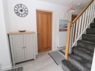 A hallway with a wooden door a grey cabinet with a bowl a round wall clock stairs with a carpet and a painting on the wall at Summer Rose in Craster