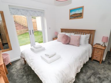 A bedroom with a wooden bed white bedding pink pillows bedside tables with lamps and glass doors opening to a garden at Summer Rose in Craster