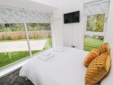 A bedroom with a bed and mustard pillows facing a wall-mounted TV with large windows showing a garden at Summer Rose in Craster